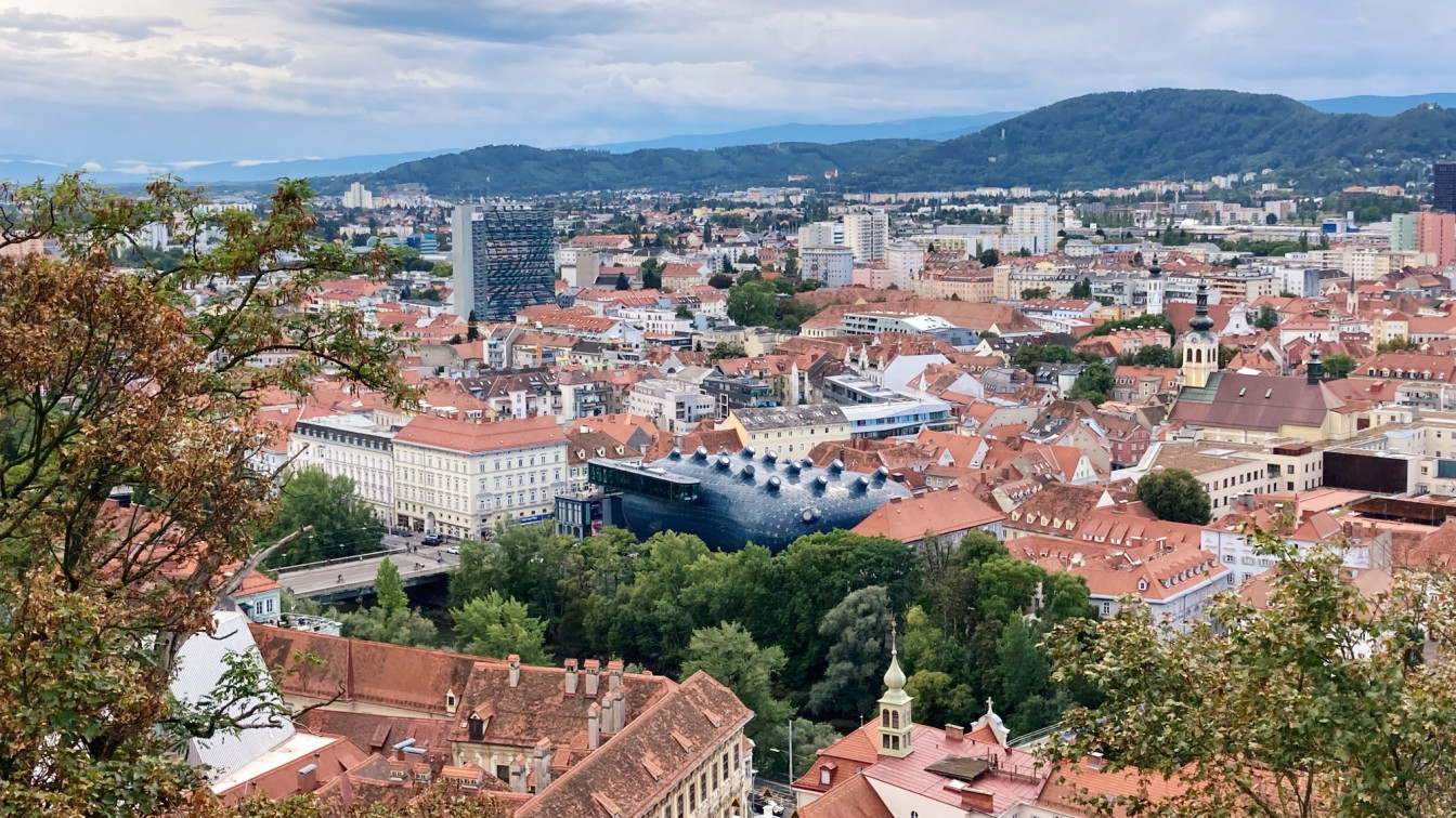Panoramablick über das Stadtzentrum von Graz mit roten Ziegeldächern, dem modernen Kunsthaus mit seiner markanten blauen Fassade in der Bildmitte und einer Mischung aus historischen und modernen Gebäuden. Im Hintergrund erstrecken sich sanfte Hügel unter bewölktem Himmel, im Vordergrund rahmen Bäume den Ausblick ein