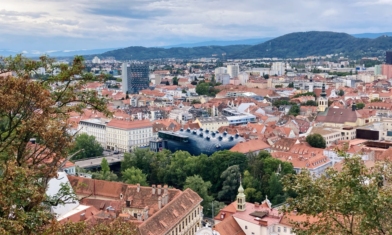 Panoramablick über das Stadtzentrum von Graz mit roten Ziegeldächern, dem modernen Kunsthaus mit seiner markanten blauen Fassade in der Bildmitte und einer Mischung aus historischen und modernen Gebäuden. Im Hintergrund erstrecken sich sanfte Hügel unter bewölktem Himmel, im Vordergrund rahmen Bäume den Ausblick ein