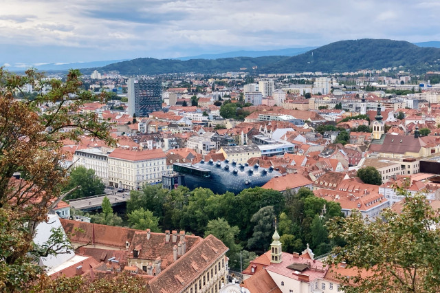 Panoramablick über das Stadtzentrum von Graz mit roten Ziegeldächern, dem modernen Kunsthaus mit seiner markanten blauen Fassade in der Bildmitte und einer Mischung aus historischen und modernen Gebäuden. Im Hintergrund erstrecken sich sanfte Hügel unter bewölktem Himmel, im Vordergrund rahmen Bäume den Ausblick ein