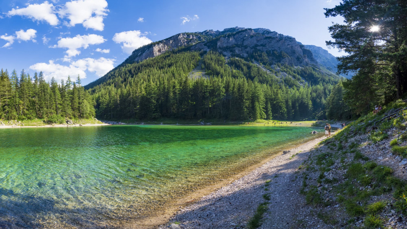 Kristallklarer Grüner See mit türkisgrünem Wasser, umgeben von einem dichten Nadelwald und steilen Felswänden. Im Hintergrund erhebt sich ein bewaldeter Berg unter blauem Himmel mit weißen Wolken. Rechts führt ein schmaler Schotterweg am Ufer entlang, einzelne Spaziergänger sind zu erkennen.