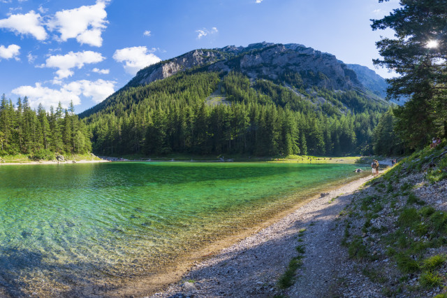 Kristallklarer Grüner See mit türkisgrünem Wasser, umgeben von einem dichten Nadelwald und steilen Felswänden. Im Hintergrund erhebt sich ein bewaldeter Berg unter blauem Himmel mit weißen Wolken. Rechts führt ein schmaler Schotterweg am Ufer entlang, einzelne Spaziergänger sind zu erkennen.