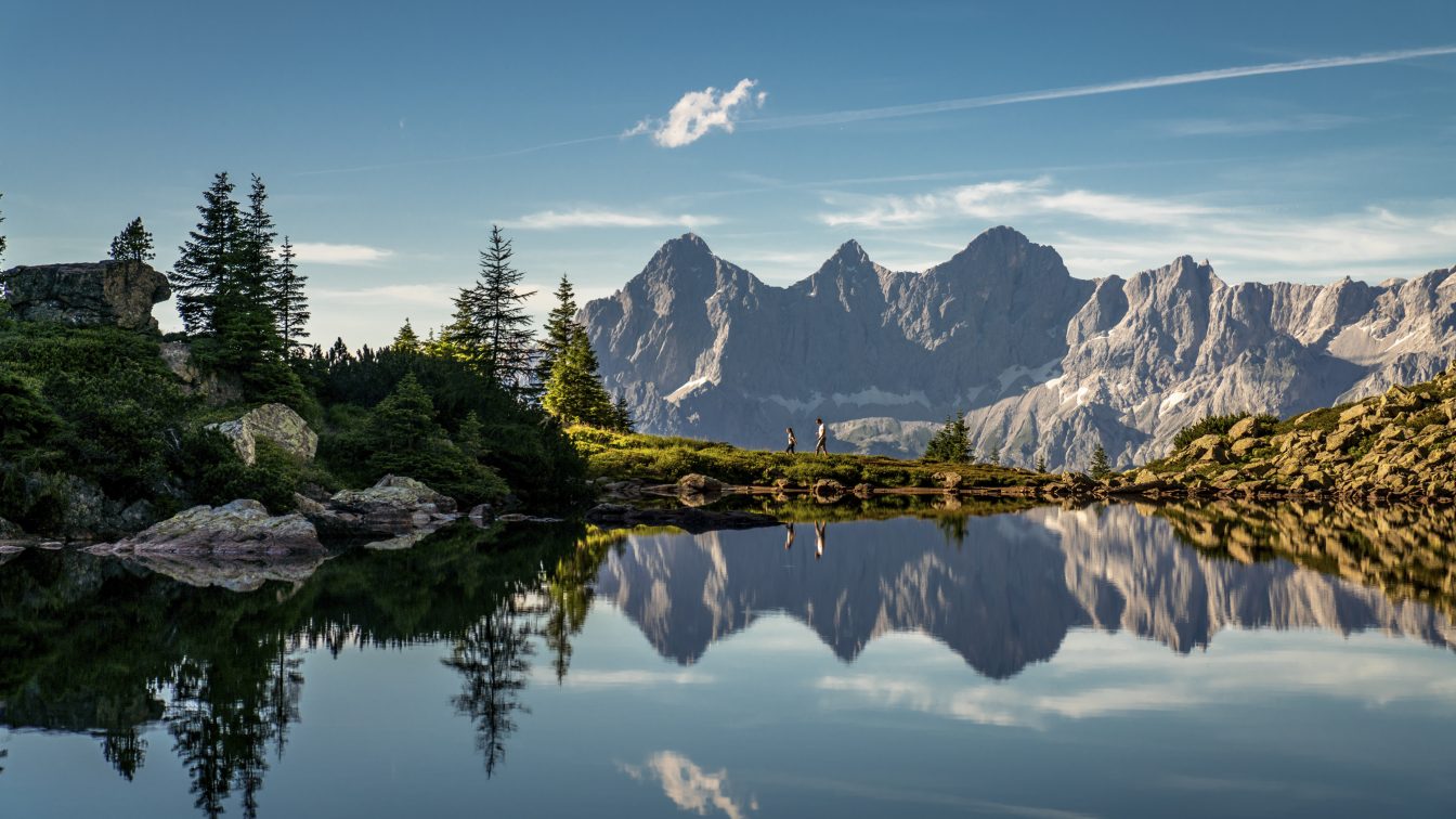 Glasklarer Bergsee mit perfekter Spiegelung der markanten Dachstein-Bergkette im Wasser. Im Vordergrund umrahmen Tannen, Felsen und Almwiesen den ruhigen See. Die Szenerie liegt unter klarem Himmel in der Morgen- oder Abendsonne, die das felsige Panorama in warmes Licht taucht.
