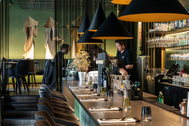 Stilvoll eingerichteter Innenraum des Restaurants Vina in Graz. Im Vordergrund eine elegante Bar mit gedeckten Plätzen, Flaschen und Gläsern. Schwarze, konisch geformte Lampen hängen über der Theke. Im Hintergrund bedienen Mitarbeitende an der Bar, während Gäste an Tischen im erhöhten Sitzbereich sitzen. Die grüne Wand mit großem Wandgemälde zweier stilisierter Figuren in traditionellen vietnamesischen Kleidern verleiht dem Raum ein besonderes Ambiente.