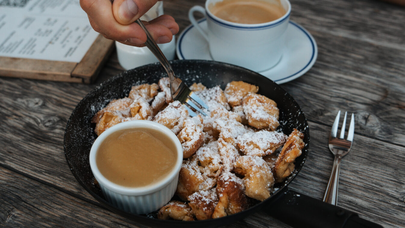 Im Bild wird eine Hand gezeigt, die mit einer Gabel Kaiserschmarrn aus einer Pfanne isst, der mit Puderzucker bestäubt ist. Daneben steht ein kleines Schälchen mit Apfelmus sowie eine Tasse Kaffee auf einem Holztisch.