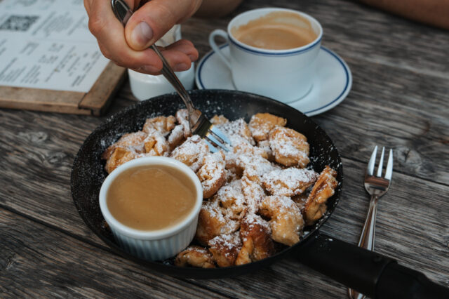 Im Bild wird eine Hand gezeigt, die mit einer Gabel Kaiserschmarrn aus einer Pfanne isst, der mit Puderzucker bestäubt ist. Daneben steht ein kleines Schälchen mit Apfelmus sowie eine Tasse Kaffee auf einem Holztisch.