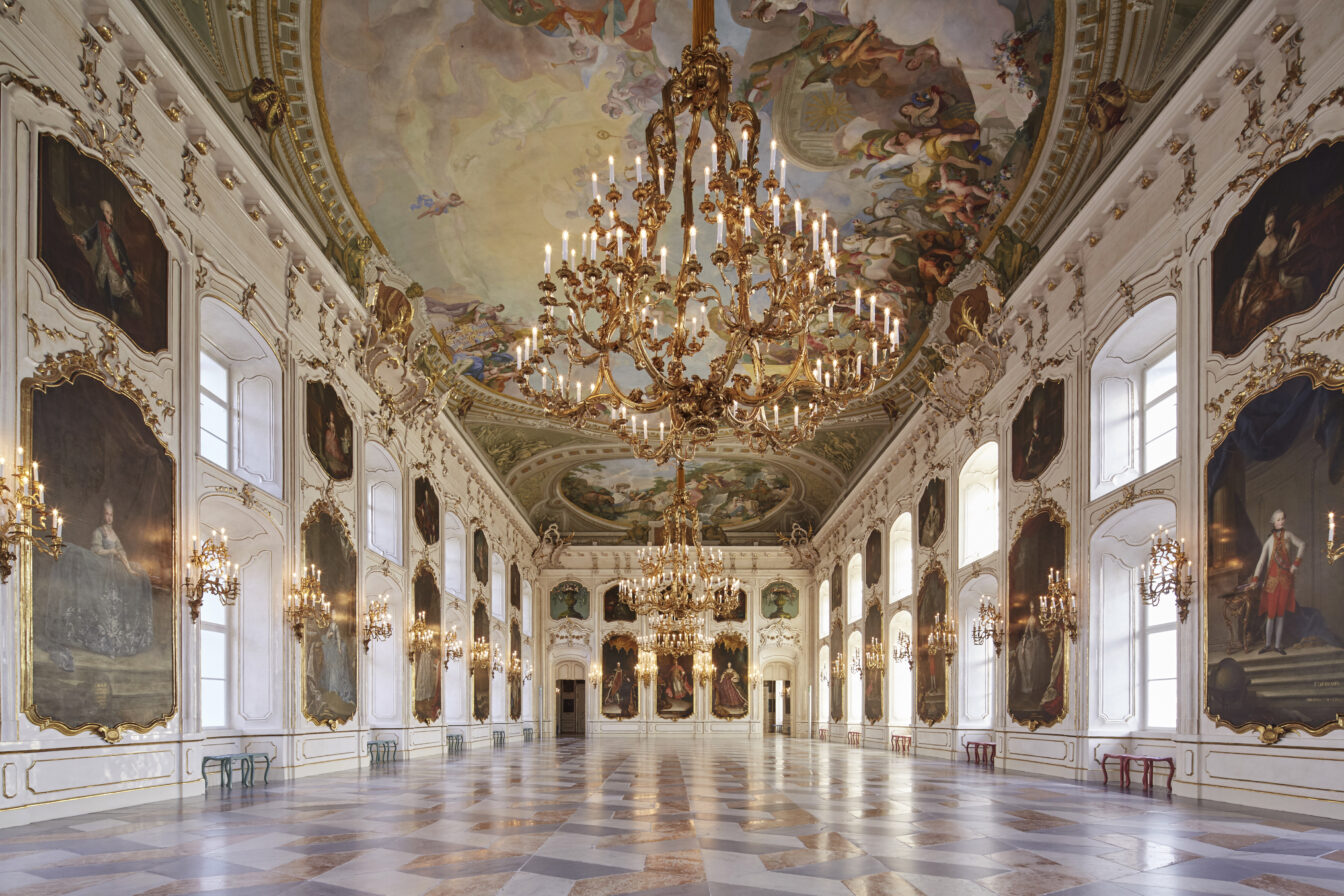 Das Bild zeigt den prächtigen Riesensaal der Kaiserlichen Hofburg in Innsbruck. Der langgestreckte Saal ist reich verziert mit prunkvollen, goldenen Kronleuchtern, die von der Decke hängen, und großformatigen Gemälden historischer Persönlichkeiten an den Wänden. Die aufwendig bemalte Decke zeigt detailreiche Szenen in Pastellfarben. Der Saal ist lichtdurchflutet, mit großen Fenstern an den Seiten, die dem Raum eine majestätische Atmosphäre verleihen. Der Marmorboden und die vergoldeten Verzierungen unterstreichen den barocken Stil und die historische Bedeutung dieses prunkvollen Festsaals.