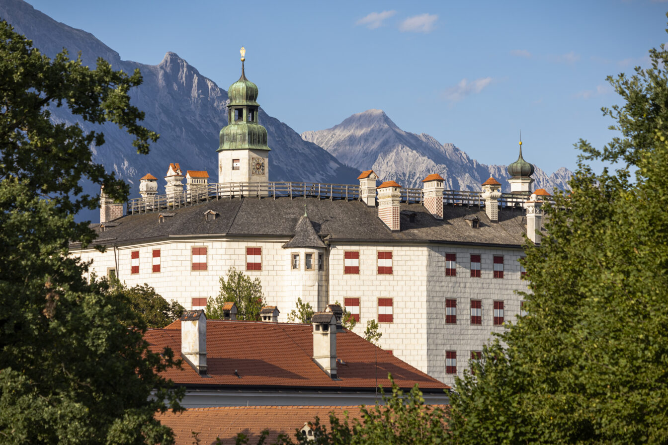 Das Bild zeigt Schloss Ambras in Innsbruck, umgeben von üppigen Bäumen und vor der Kulisse der imposanten Alpen. Das Schloss ist bekannt für seine helle Fassade mit roten Fensterläden und seine charakteristischen Türmchen. Die grüne Kuppel des Hauptturms ragt prominent hervor. Der strahlend blaue Himmel und die mächtigen Berge im Hintergrund betonen die malerische Lage des Schlosses. Es vermittelt ein Gefühl von historischer Bedeutung und natürlicher Schönheit, eingebettet in die alpine Landschaft Tirols.