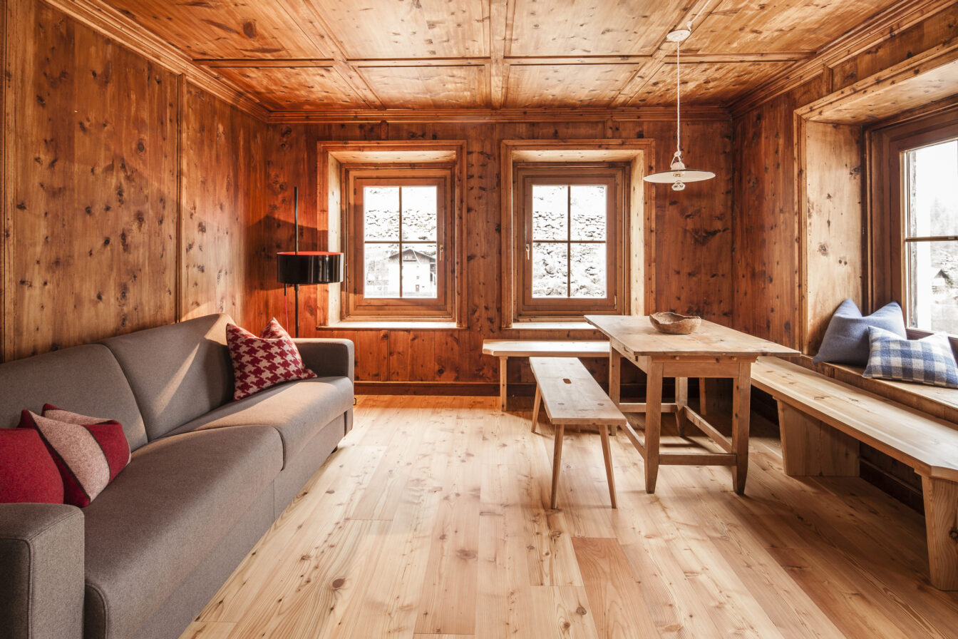 Gemütliche Alpenlodge in Südtirol mit warmen Holzwänden, grauem Sofa und roten Kissen, rustikalem Esstisch mit Bänken und großen Fenstern, die einen Blick auf die schneebedeckten Berge bieten.