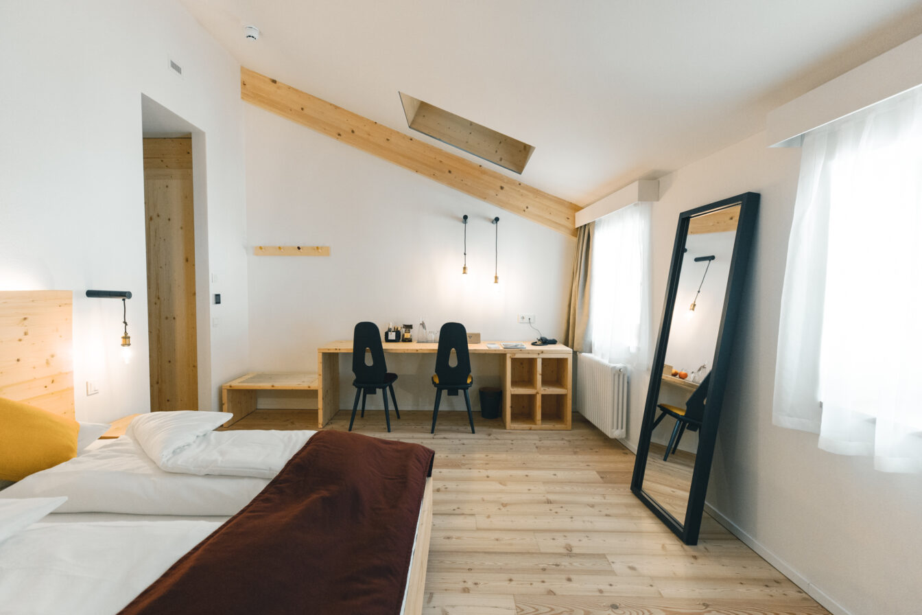 Ein minimalistisches Hotelzimmer im Gasthof zum Hirschen, gestaltet mit viel Holz. Es gibt zwei Stühle vor einem Schreibtisch mit einer Lampe und einem großen Spiegel, der an der Wand steht. Der Raum ist hell und freundlich, mit einem schrägen Holzbalken an der Decke und großen Fenstern, durch die Tageslicht einfällt. Ein Bett mit weißer Bettwäsche und einer braunen Decke steht links im Bild.