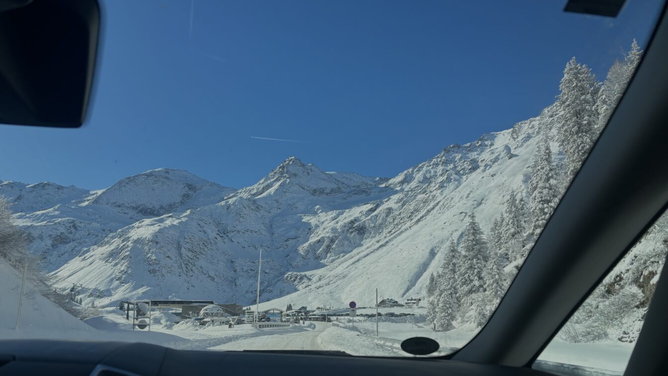 Das Bild zeigt eine beeindruckende Berglandschaft, aufgenommen aus einem Auto heraus. Im Vordergrund ist die Windschutzscheibe des Fahrzeugs sichtbar, während sich die schneebedeckten Berge und Bäume im Hintergrund erstrecken. Der Himmel ist klar und strahlend blau, was einen schönen Kontrast zu der weißen Schneedecke bildet. Es scheint eine winterliche Bergstraße zu sein, mit einem Gebäude und einigen Fahrzeugen im Talbereich. Das Bild vermittelt eine ruhige und majestätische Atmosphäre eines Bergortes.