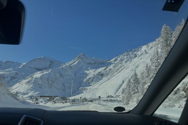 Das Bild zeigt eine beeindruckende Berglandschaft, aufgenommen aus einem Auto heraus. Im Vordergrund ist die Windschutzscheibe des Fahrzeugs sichtbar, während sich die schneebedeckten Berge und Bäume im Hintergrund erstrecken. Der Himmel ist klar und strahlend blau, was einen schönen Kontrast zu der weißen Schneedecke bildet. Es scheint eine winterliche Bergstraße zu sein, mit einem Gebäude und einigen Fahrzeugen im Talbereich. Das Bild vermittelt eine ruhige und majestätische Atmosphäre eines Bergortes.