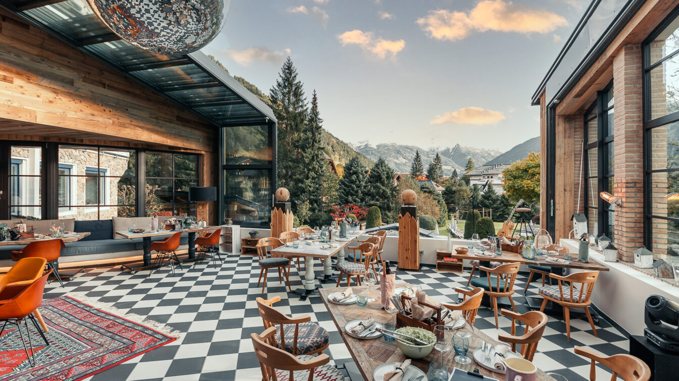 Sendlhofers in Salzburg. Moderner Wintergarten eines Restaurants mit großen Fensterfronten und Bergpanorama im Hintergrund. Der Raum wirkt gemütlich und einladend, mit Holzmöbeln, kariertem Boden und einer großen Diskokugel an der Decke. Der Blick reicht über grüne Nadelbäume und Berge unter blauem Himmel mit wenigen Wolken.