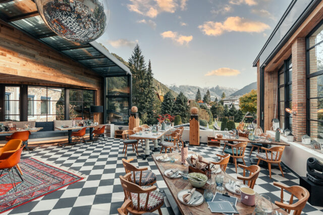 Sendlhofers in Salzburg. Moderner Wintergarten eines Restaurants mit großen Fensterfronten und Bergpanorama im Hintergrund. Der Raum wirkt gemütlich und einladend, mit Holzmöbeln, kariertem Boden und einer großen Diskokugel an der Decke. Der Blick reicht über grüne Nadelbäume und Berge unter blauem Himmel mit wenigen Wolken.