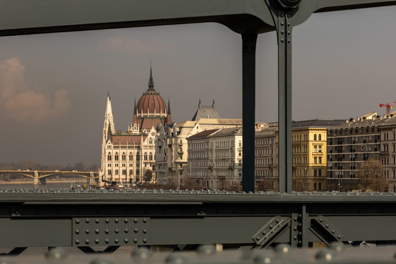 Blick durch die Stahlkonstruktion der Széchenyi-Kettenbrücke auf das ungarische Parlamentsgebäude in Budapest. Die imposante Kuppel und neugotischen Türme des Parlaments strahlen in warmem Licht, flankiert von eleganten Altbauten entlang des Donauufers. Im Hintergrund ist die Margaretenbrücke zu erkennen. Die Szene zeigt eines der bekanntesten Highlights und Fotomotive in Budapest aus ungewöhnlicher Perspektive.