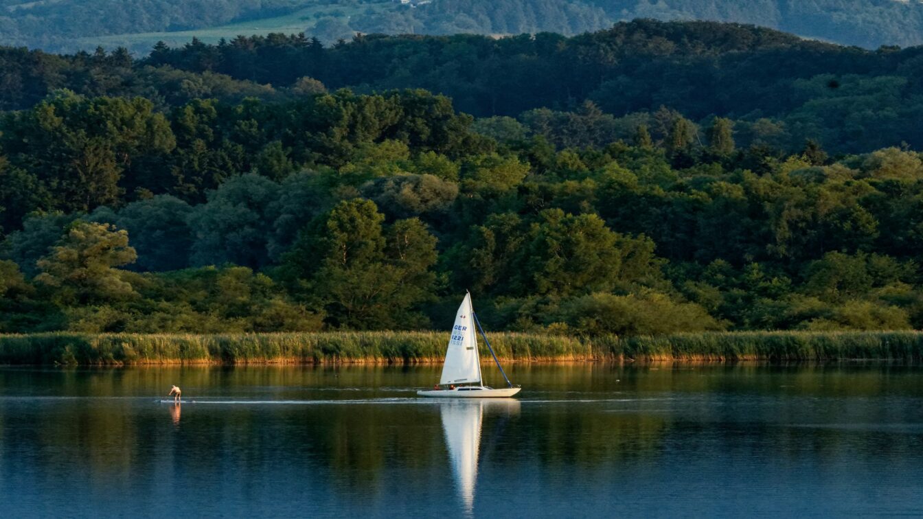 Segelboot auf dem Chiemsee bei Sonnenuntergang vor bewaldeter Hügellandschaft in Bayern