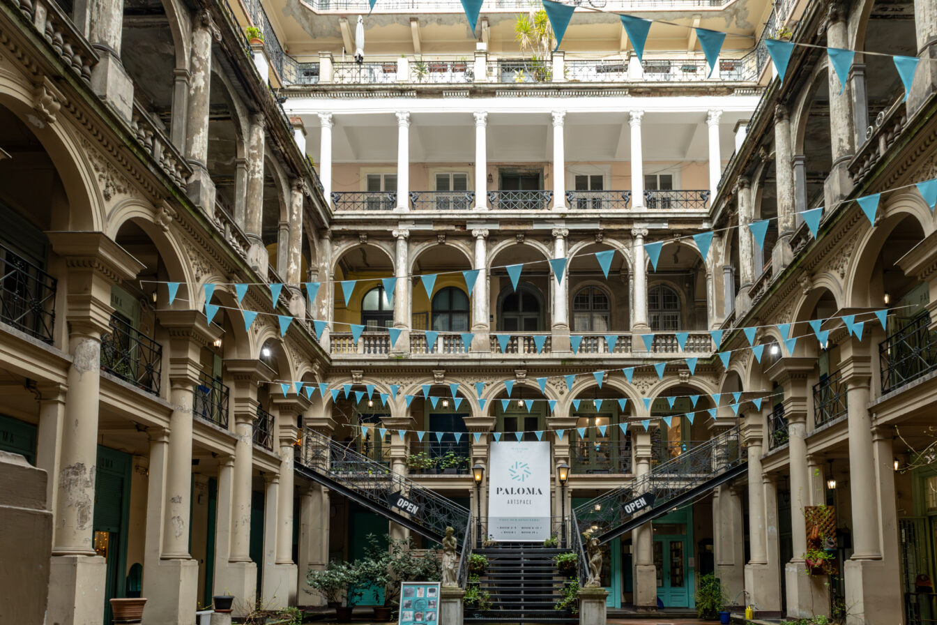 Innenhof des Paloma Artspace in Budapest mit historischem Säulengang, verspielter Architektur und blauen Wimpelgirlanden. Der kreative Kunst- und Design-Hotspot beherbergt Ateliers lokaler Künstlerinnen und Designerinnen in einem charmant gealterten Altbau. Der zentrale Eingang mit Treppe und dem Paloma-Schild lädt Besucher*innen zum Entdecken ein – ein echter Geheimtipp für Kunst- und Kulturfans in Budapest.