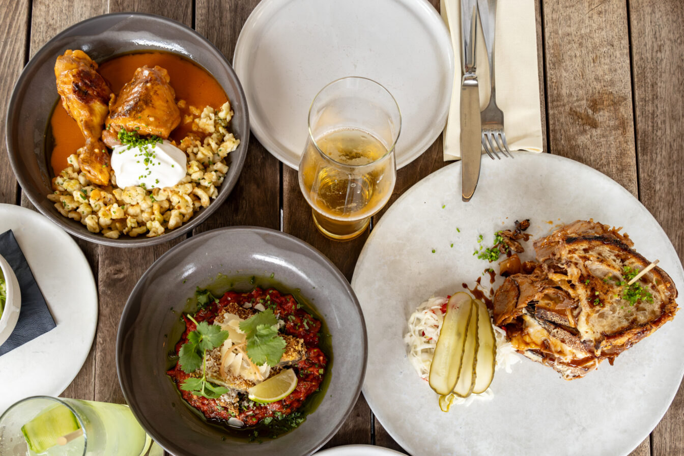 Tisch mit ungarischen Spezialitäten im Restaurant BESTIA Budapest. Zu sehen sind ein Paprikahendl mit Nockerl und Sauerrahm, ein Gericht mit gegrilltem Gemüse und Kräutern sowie ein Pulled Pork Sandwich mit Sauerkraut und Essiggurke. Dazu ein Glas Bier und frische Limonade – angerichtet auf rustikalem Holztisch. Das BESTIA ist bekannt für moderne ungarische Küche, hochwertige Zutaten und entspannte Atmosphäre nahe der Basilika.