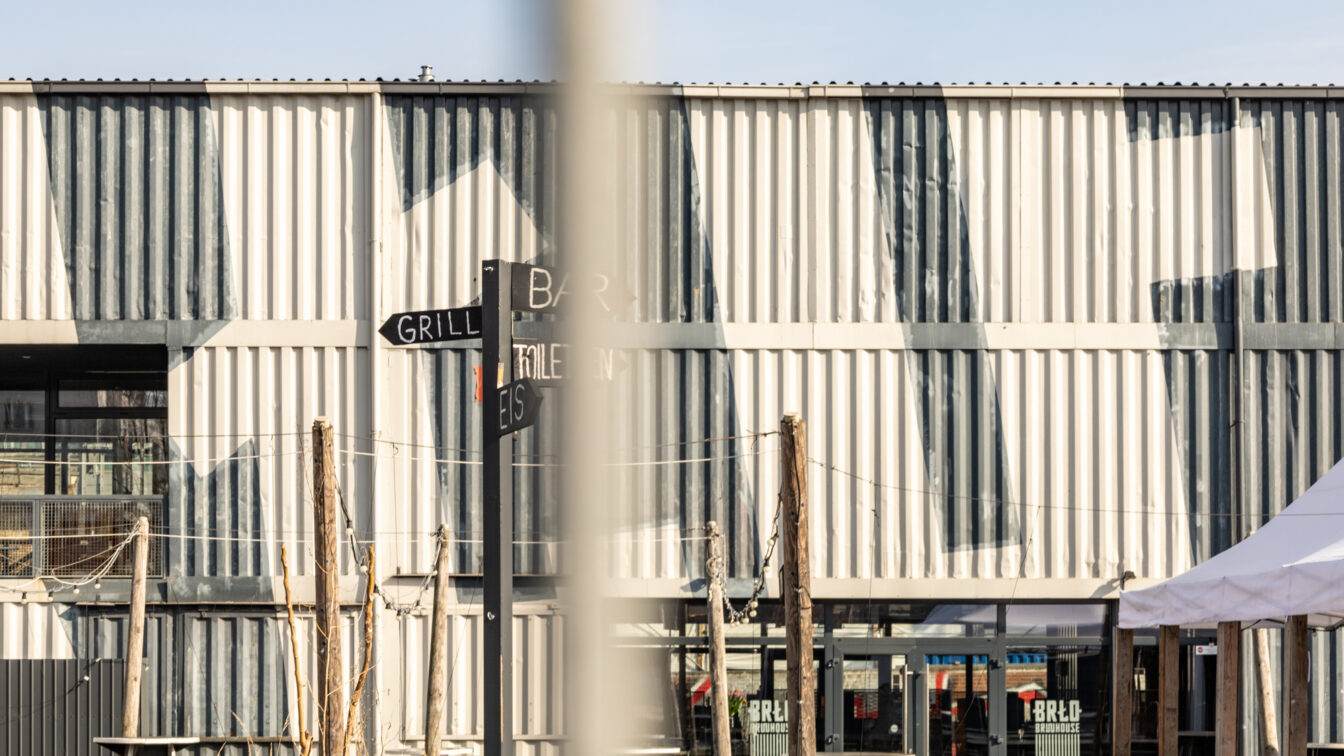 Außenansicht des BRLO Brwhouse Biergartens in Berlin mit einer modernen, industriellen Ästhetik. Die Fassade besteht aus weiß-grau gestrichenen Schiffscontainern mit geometrischen Mustern. Im Vordergrund steht ein schwarzer Wegweiser mit den Beschriftungen 'Grill', 'Bar', 'Eis' und 'Toiletten'. Mehrere Holztische und Sitzgelegenheiten sind über das Gelände verteilt, während Lichterketten zwischen Holzpfosten gespannt sind. Auf der rechten Seite ist ein weißes Zelt zu sehen, das zum Außenbereich des Biergartens gehört. Die Szene wird von hellem Sonnenlicht beleuchtet, das starke Schatten auf die Wand wirft.