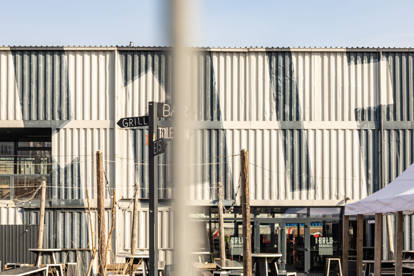 Außenansicht des BRLO Brwhouse Biergartens in Berlin mit einer modernen, industriellen Ästhetik. Die Fassade besteht aus weiß-grau gestrichenen Schiffscontainern mit geometrischen Mustern. Im Vordergrund steht ein schwarzer Wegweiser mit den Beschriftungen 'Grill', 'Bar', 'Eis' und 'Toiletten'. Mehrere Holztische und Sitzgelegenheiten sind über das Gelände verteilt, während Lichterketten zwischen Holzpfosten gespannt sind. Auf der rechten Seite ist ein weißes Zelt zu sehen, das zum Außenbereich des Biergartens gehört. Die Szene wird von hellem Sonnenlicht beleuchtet, das starke Schatten auf die Wand wirft.