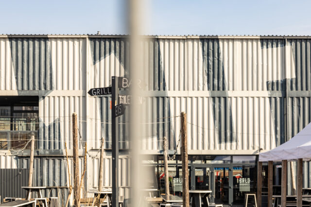 Außenansicht des BRLO Brwhouse Biergartens in Berlin mit einer modernen, industriellen Ästhetik. Die Fassade besteht aus weiß-grau gestrichenen Schiffscontainern mit geometrischen Mustern. Im Vordergrund steht ein schwarzer Wegweiser mit den Beschriftungen 'Grill', 'Bar', 'Eis' und 'Toiletten'. Mehrere Holztische und Sitzgelegenheiten sind über das Gelände verteilt, während Lichterketten zwischen Holzpfosten gespannt sind. Auf der rechten Seite ist ein weißes Zelt zu sehen, das zum Außenbereich des Biergartens gehört. Die Szene wird von hellem Sonnenlicht beleuchtet, das starke Schatten auf die Wand wirft.