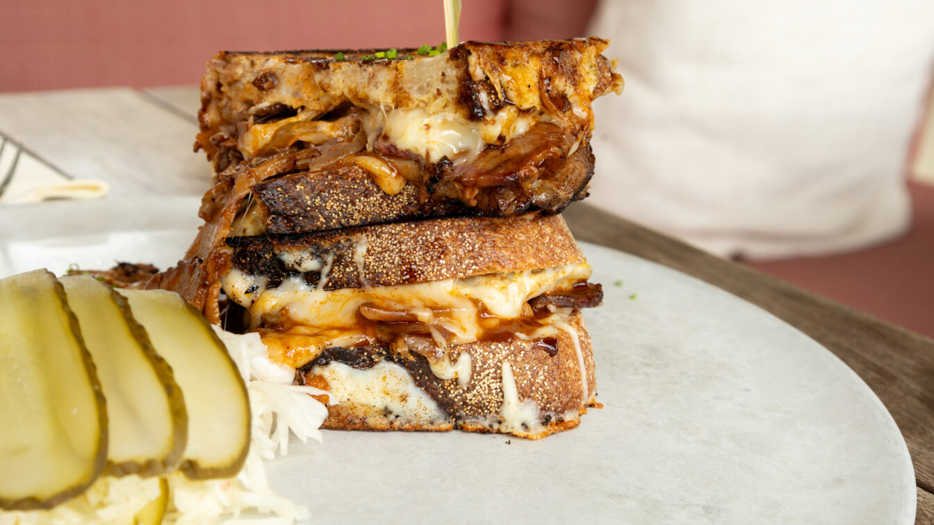 Nahaufnahme eines herzhaften Grilled Cheese Sandwiches mit geschmolzenem Käse und Pulled Pork, serviert auf einem hellen Teller mit Sauerkraut und Gurkenscheiben. Das Gericht steht auf einem Holztisch in gemütlicher Restaurantatmosphäre in Budapest. Perfekt für Foodies auf der Suche nach den besten Bars und Restaurants in Budapest mit moderner, kreativer Küche und Wohlfühlfaktor.