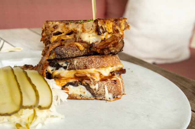 Nahaufnahme eines herzhaften Grilled Cheese Sandwiches mit geschmolzenem Käse und Pulled Pork, serviert auf einem hellen Teller mit Sauerkraut und Gurkenscheiben. Das Gericht steht auf einem Holztisch in gemütlicher Restaurantatmosphäre in Budapest. Perfekt für Foodies auf der Suche nach den besten Bars und Restaurants in Budapest mit moderner, kreativer Küche und Wohlfühlfaktor.
