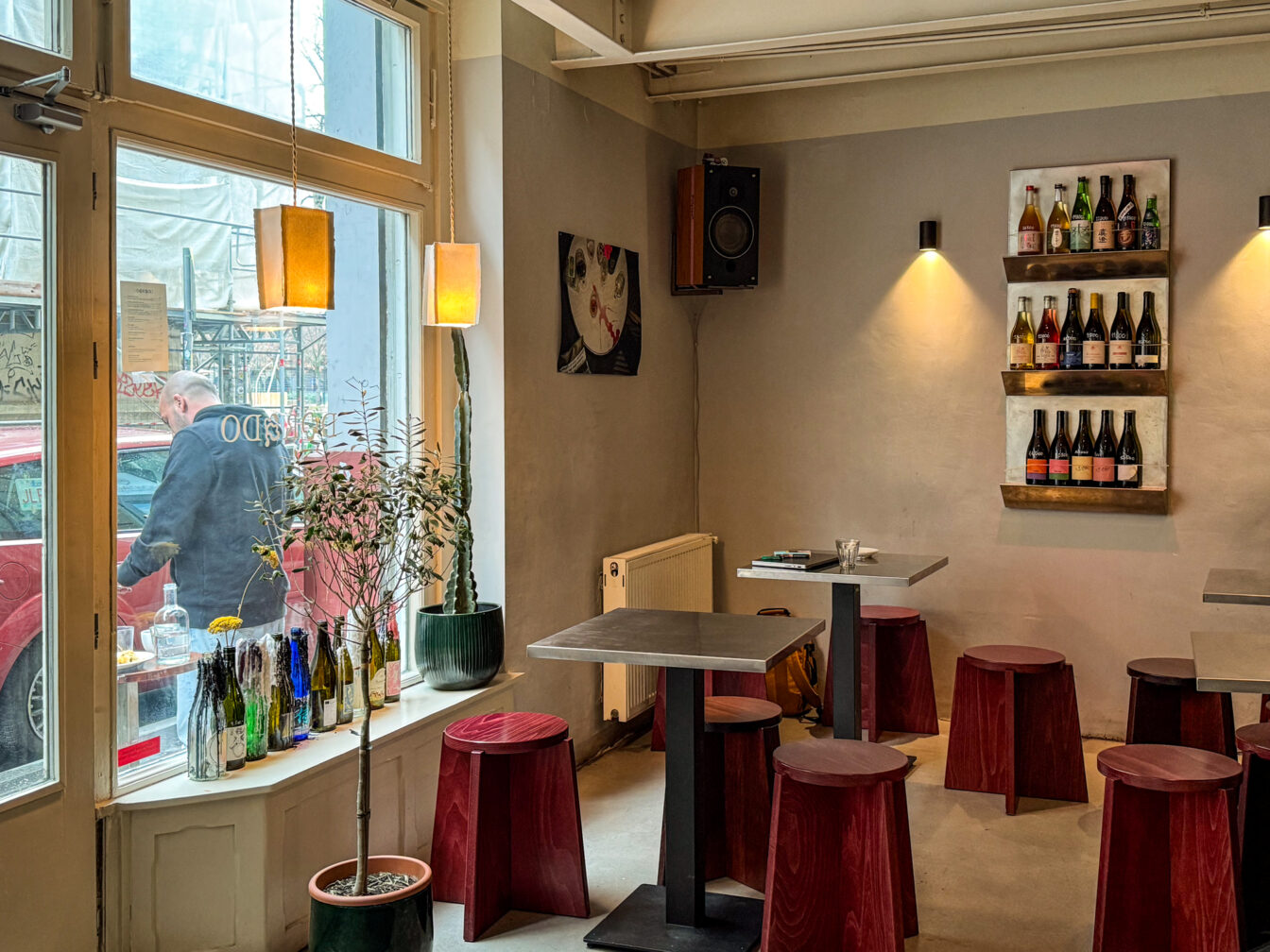 Innenaufnahme des minimalistischen Dorado Cafés in Budapest mit klaren Linien, schlichten Holzhockern und modernen Tischen. An der Wand hängt ein Regal mit Naturweinen, während sanftes Licht eine gemütliche Atmosphäre schafft. Auf der Fensterbank stehen bunte Flaschen und Pflanzen, draußen vor dem Fenster ist ein Gast zu sehen. Das Dorado ist bekannt für Specialty Coffee, Naturwein und entspanntes Design im Herzen des VII. Bezirks.