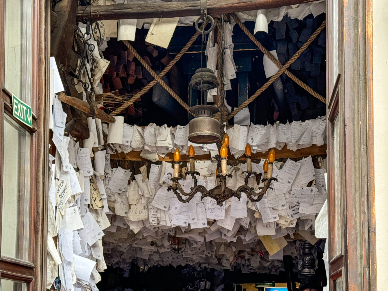 Blick ins Innere des For Sale Pub in Budapest, wo hunderte handgeschriebene Notizen, Zettel und Botschaften die Decke und Wände bedecken. In der Mitte hängt ein rustikaler Kronleuchter unter Seilen und Laternen – umgeben von kreativem Chaos. Das einzigartige Pub ist bekannt für seine besondere Atmosphäre, bei der Gäste persönliche Nachrichten hinterlassen und Teil des Ortes werden.