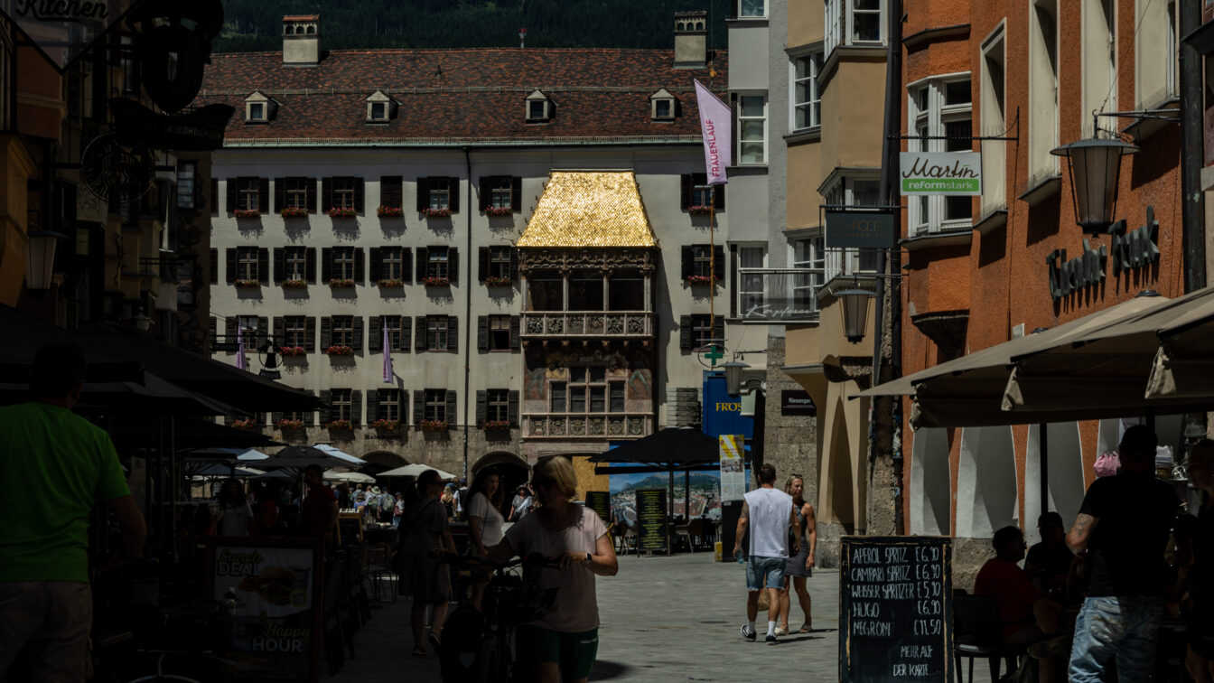 Eine belebte Fußgängerzone in Innsbruck, Österreich, mit Menschen, die in Straßencafés sitzen oder spazieren gehen. Im Hintergrund befindet sich das berühmte „Goldene Dachl“, ein spätgotischer Erker mit einem Dach aus goldenen Schindeln, an der Fassade eines historischen Gebäudes. Rechts und links der Straße stehen bunte, mehrstöckige Häuser mit Läden und Restaurants. Im Vordergrund schiebt eine Frau ein Fahrrad, während andere Besucher die Szene genießen. Im Hintergrund sind grüne Berge sichtbar.