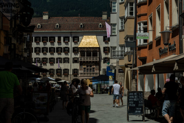 Eine belebte Fußgängerzone in Innsbruck, Österreich, mit Menschen, die in Straßencafés sitzen oder spazieren gehen. Im Hintergrund befindet sich das berühmte „Goldene Dachl“, ein spätgotischer Erker mit einem Dach aus goldenen Schindeln, an der Fassade eines historischen Gebäudes. Rechts und links der Straße stehen bunte, mehrstöckige Häuser mit Läden und Restaurants. Im Vordergrund schiebt eine Frau ein Fahrrad, während andere Besucher die Szene genießen. Im Hintergrund sind grüne Berge sichtbar.