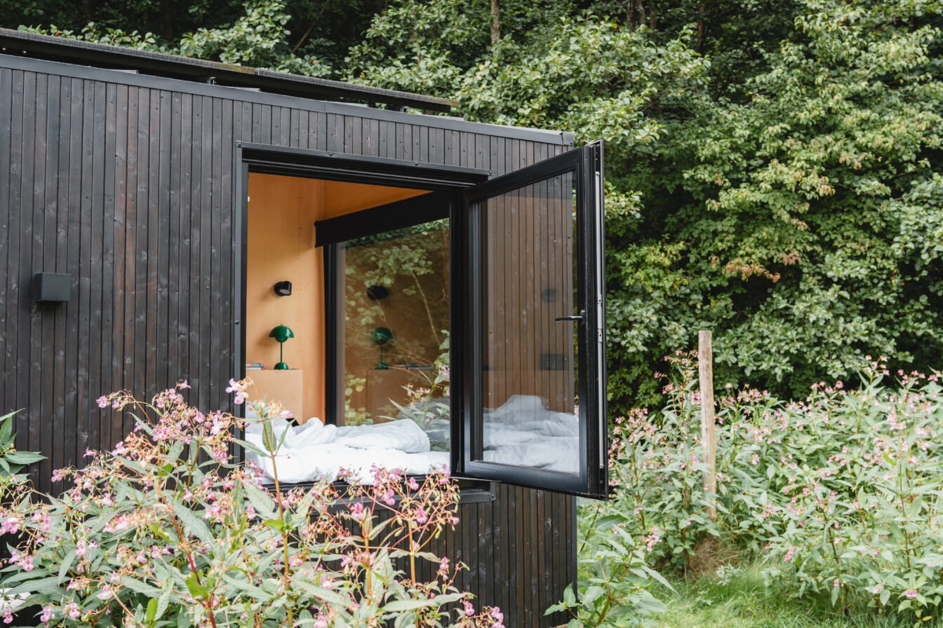 Modernes RAUS Tiny House Cabin mit geöffnetem Panoramafenster im Grünen, minimalistisches Design mit Holzverkleidung und Blick ins Schlafzimmer.
