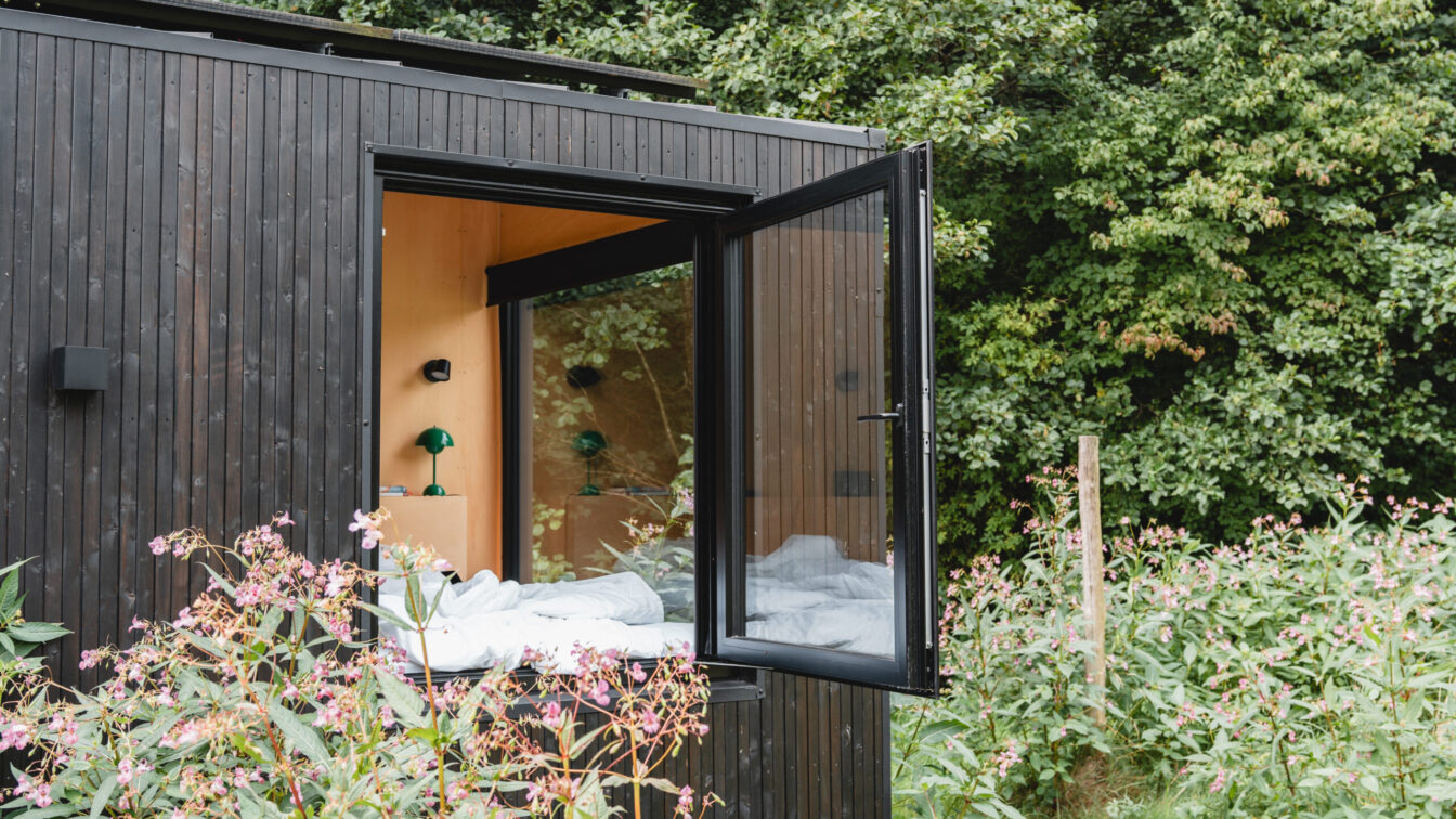 Modernes RAUS Tiny House Cabin mit geöffnetem Panoramafenster im Grünen, minimalistisches Design mit Holzverkleidung und Blick ins Schlafzimmer.