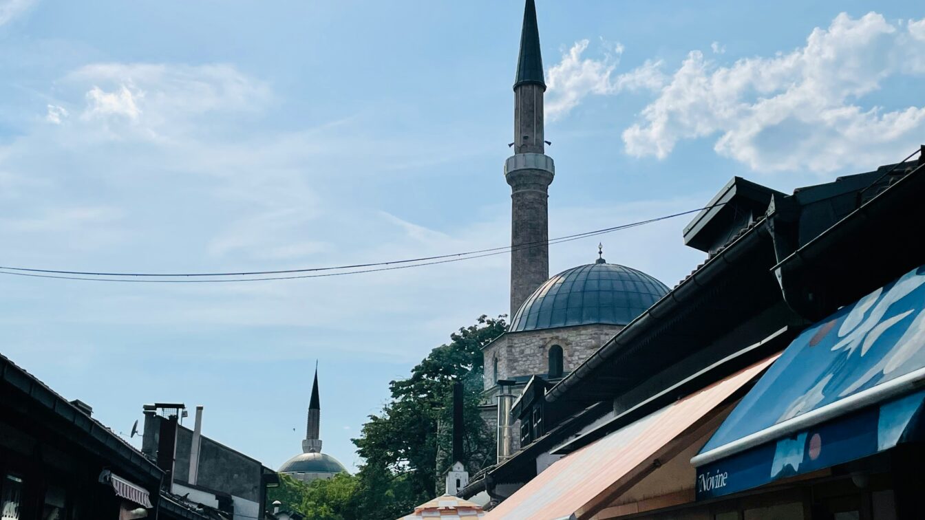 Belebte Gasse mit Restaurants und Cafés in der Altstadt von Sarajevo bei sonnigem Wetter. Im Hintergrund sind zwei Moscheen mit markanten Minaretten und Kuppeln zu sehen, die sich über den Dächern und bunten Sonnenschirmen der Lokale erheben.