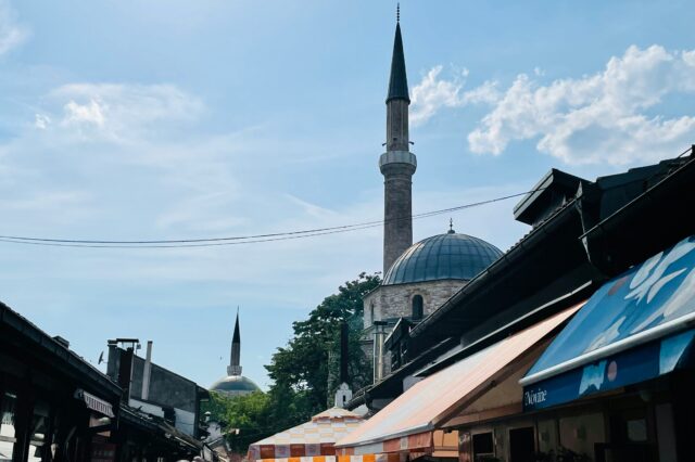 Belebte Gasse mit Restaurants und Cafés in der Altstadt von Sarajevo bei sonnigem Wetter. Im Hintergrund sind zwei Moscheen mit markanten Minaretten und Kuppeln zu sehen, die sich über den Dächern und bunten Sonnenschirmen der Lokale erheben.