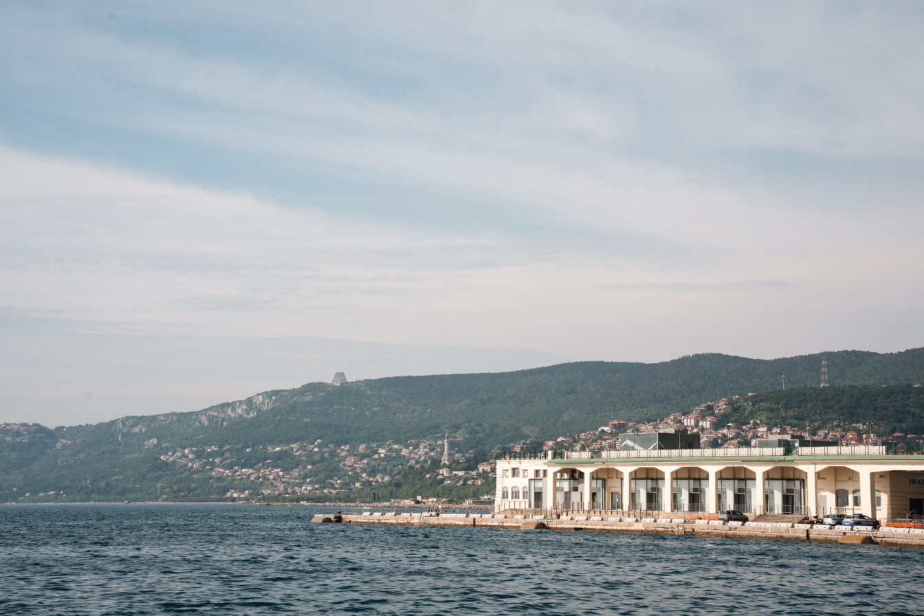 Blick auf die Küste von Triest mit dem historischen Hafenlagerhaus, azurblauem Meer und grünen Hügeln im Hintergrund – mediterrane Hafenstadt mit italienischem Charme und maritimem Flair.