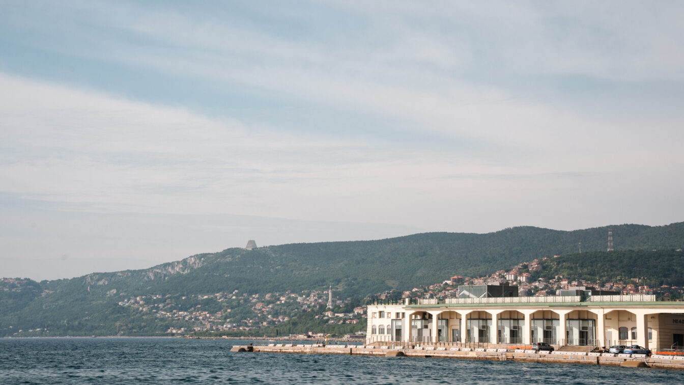 Blick auf die Küste von Triest mit dem historischen Hafenlagerhaus, azurblauem Meer und grünen Hügeln im Hintergrund – mediterrane Hafenstadt mit italienischem Charme und maritimem Flair.