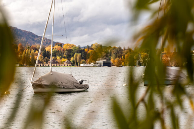 Blick durch herbstlich gefärbte Äste auf ein ruhiges Gewässer mit zwei abgedeckten Segelbooten, die vor Anker liegen. Im Hintergrund sind farbenfrohe Bäume, Gebäude am Ufer und bewaldete Hügel unter einem bewölkten Himmel zu sehen.“