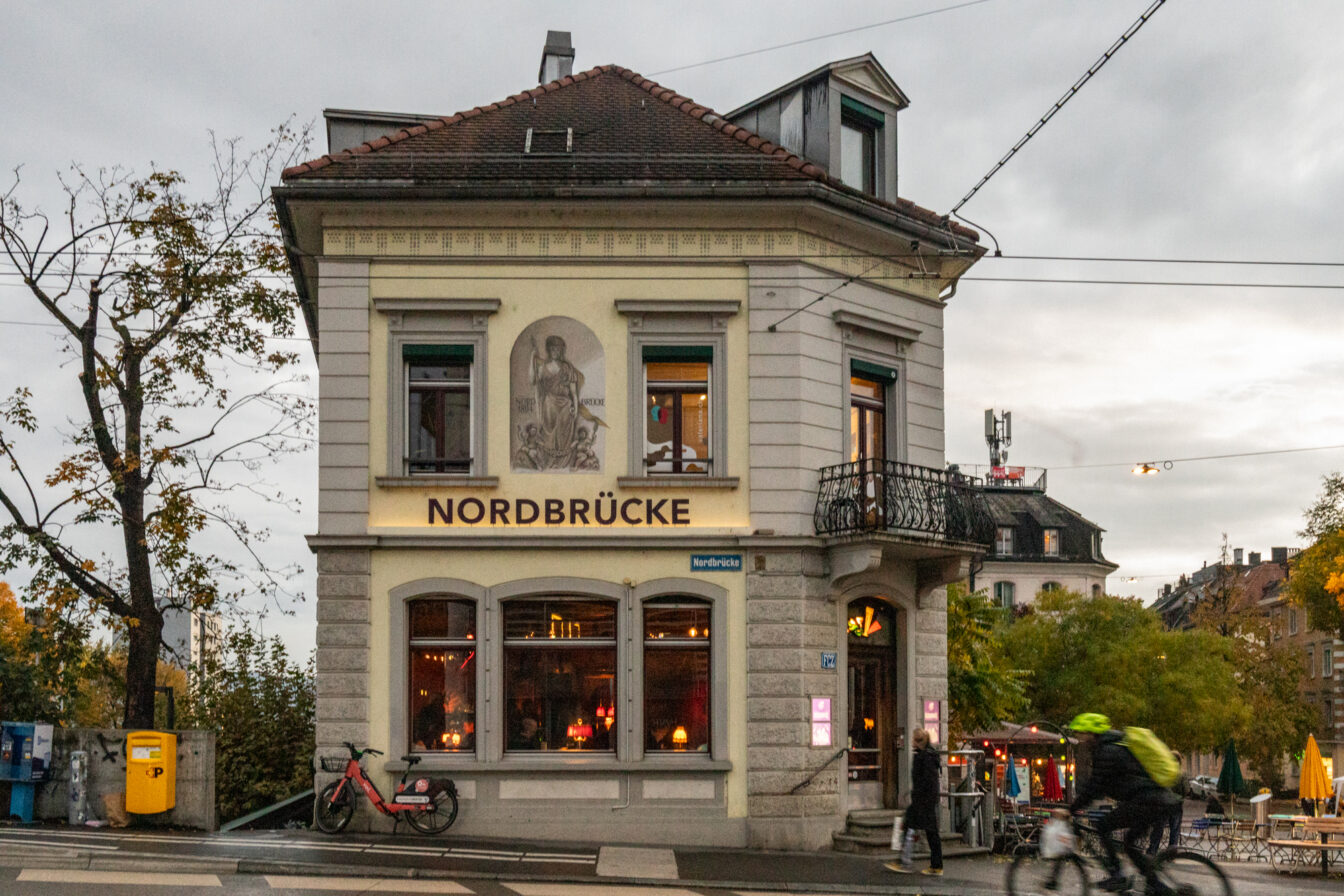 Straßenszene in Zürich mit dem Café-Restaurant Nordbrücke in einem historischen Eckgebäude. Im Vordergrund fährt ein Radfahrer vorbei, während eine Person zu Fuß das Lokal betritt. Die Umgebung ist herbstlich, mit Bäumen und Außentischen auf dem Platz neben dem Gebäude