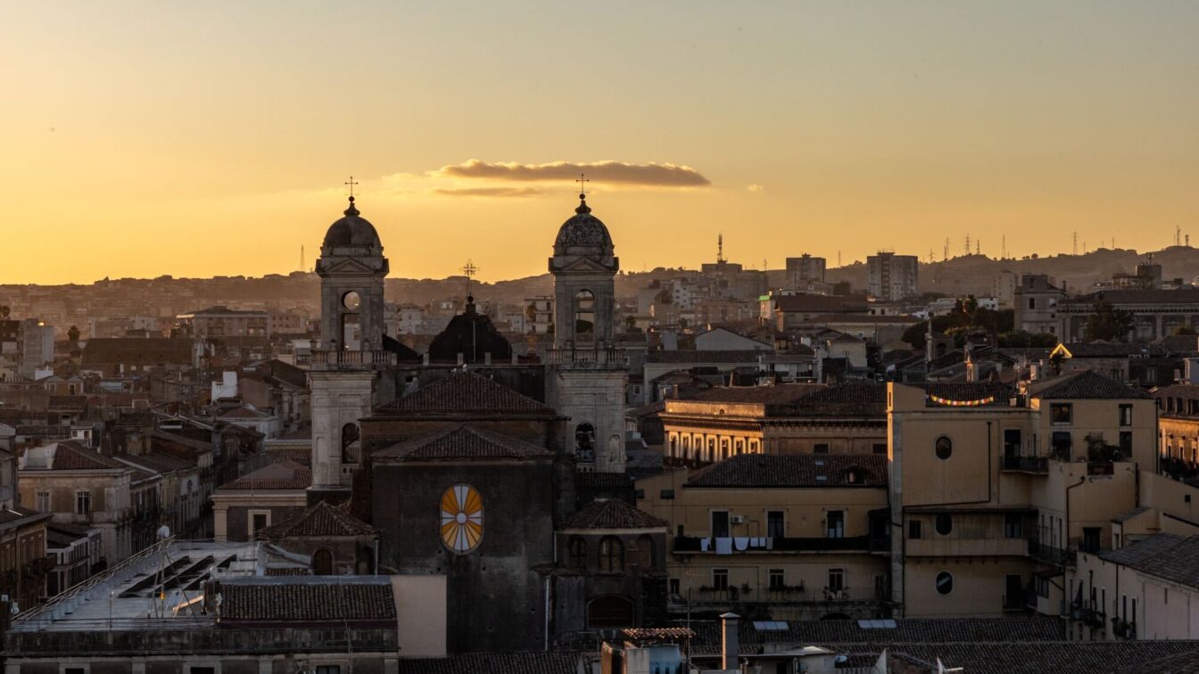 Ein Blick auf Catania, Italien, bei Sonnenuntergang. Die Silhouette zweier Kirchtürme dominiert den Vordergrund, während die Stadt im warmen Abendlicht leuchtet.