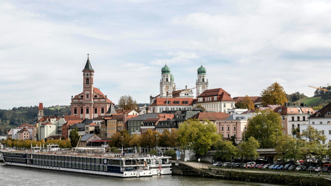 Blick auf die Altstadt von Passau mit der Donau im Vordergrund, Kreuzfahrtschiffen am Ufer, bunten Fassaden der Häuser und markanten Kirchen – links die Stadtpfarrkirche St. Paul mit hohem Turm, im Hintergrund der Dom St. Stephan mit seinen grünen Zwiebeltürmen.