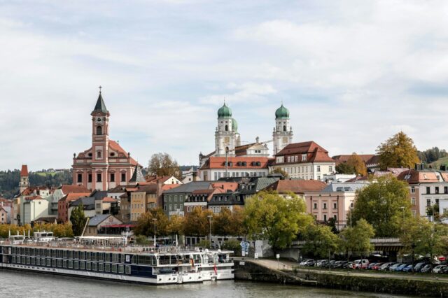 Blick auf die Altstadt von Passau mit der Donau im Vordergrund, Kreuzfahrtschiffen am Ufer, bunten Fassaden der Häuser und markanten Kirchen – links die Stadtpfarrkirche St. Paul mit hohem Turm, im Hintergrund der Dom St. Stephan mit seinen grünen Zwiebeltürmen.
