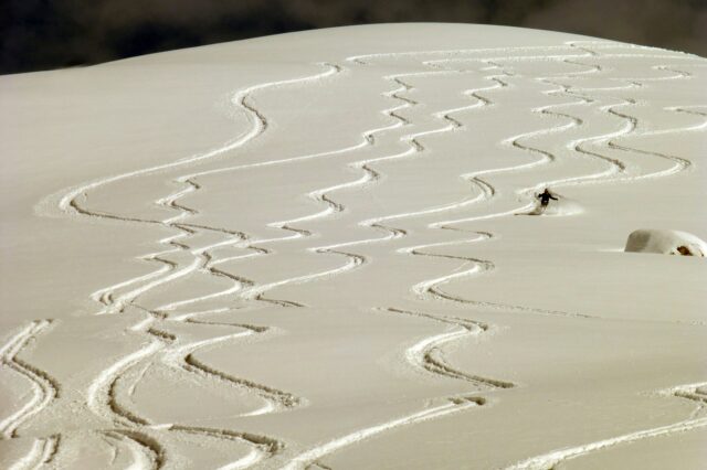Eine verschneite, unberührte Piste mit zahlreichen eleganten Zickzack-Spuren, die von Skifahrer*innen im Pulverschnee hinterlassen wurden. Eine einzelne Person fährt in der Ferne den Hang hinunter.
