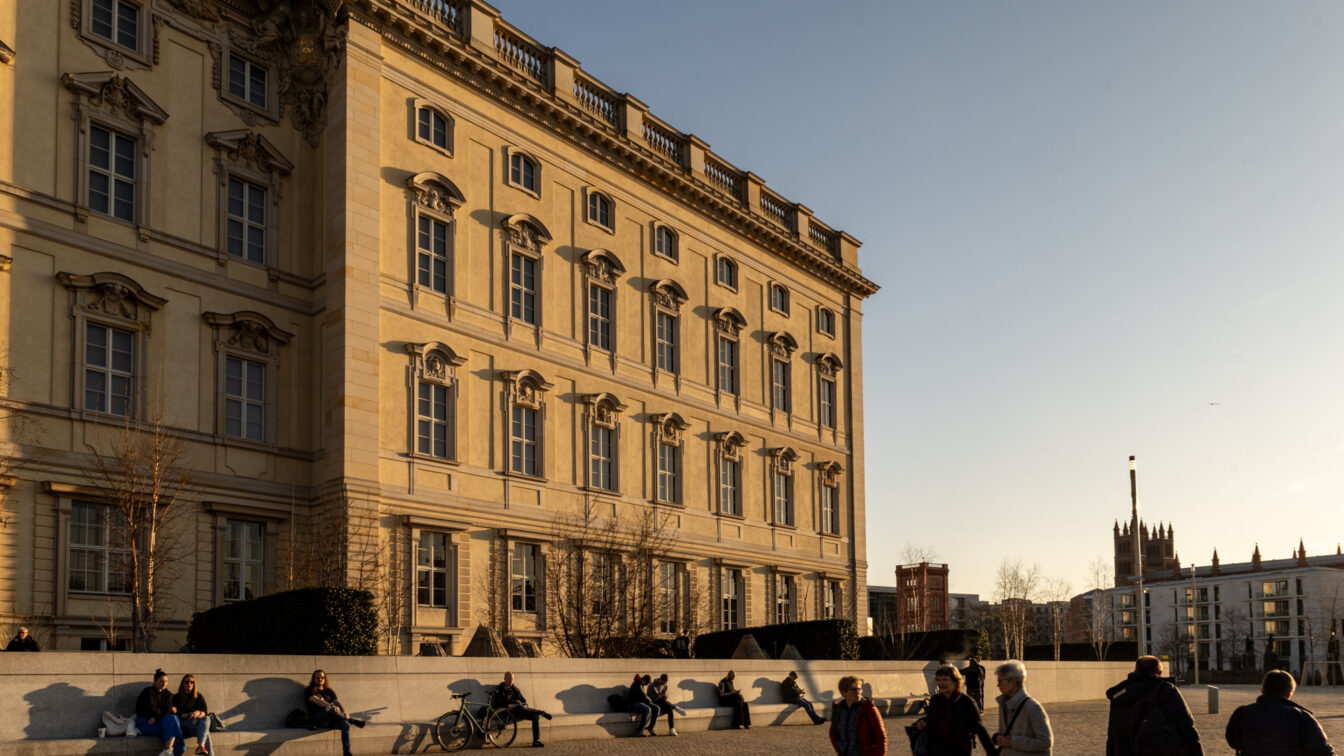 Eine historische Fassade des Berliner Schlosses im warmen Licht der tief stehenden Sonne. Der barocke Baustil mit reich verzierten Fenstern und dekorativen Elementen prägt das Gebäude. Im Vordergrund sitzen und lehnen mehrere Menschen entspannt an einer niedrigen Steinmauer, einige mit Fahrrädern. Spaziergänger bewegen sich über den weitläufigen Platz. Im Hintergrund sind moderne Gebäude und eine Kirche mit neugotischem Turm zu sehen. Die langen Schatten und das goldene Licht verleihen der Szene eine ruhige, stimmungsvolle Atmosphäre.