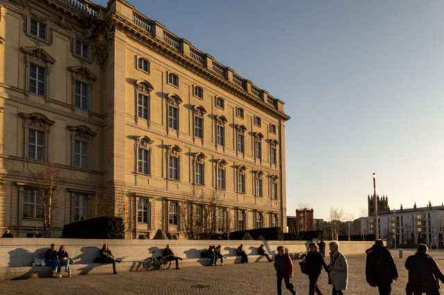 Eine historische Fassade des Berliner Schlosses im warmen Licht der tief stehenden Sonne. Der barocke Baustil mit reich verzierten Fenstern und dekorativen Elementen prägt das Gebäude. Im Vordergrund sitzen und lehnen mehrere Menschen entspannt an einer niedrigen Steinmauer, einige mit Fahrrädern. Spaziergänger bewegen sich über den weitläufigen Platz. Im Hintergrund sind moderne Gebäude und eine Kirche mit neugotischem Turm zu sehen. Die langen Schatten und das goldene Licht verleihen der Szene eine ruhige, stimmungsvolle Atmosphäre.