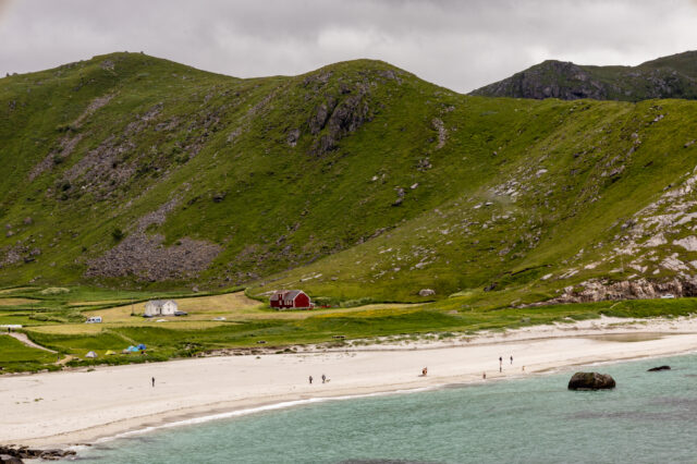 Weitläufige grüne Hügel mit Felsformationen steigen hinter einem hellen Sandstrand an. Einige kleine Häuser, darunter ein rotes Holzhaus und ein weißes Haus, liegen zwischen Wiesen am Fuß der Hügel. Menschen spazieren am Strand entlang, und das türkisfarbene Wasser trifft sanft auf die Küste unter einem bewölkten Himmel.“