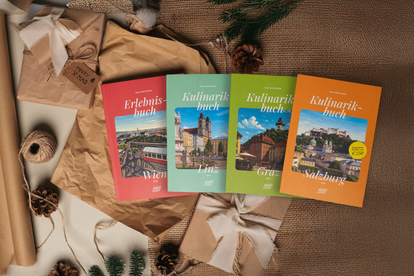 1000things Erlebnisbuch Wien sowie Kulinarikbücher für Linz, Graz und Salzburg, stimmungsvoll arrangiert mit Geschenkpapier, Tannenzweigen und Weihnachtsdeko – ideales Weihnachtsgeschenk für Kulinarik- und Erlebnisfans.