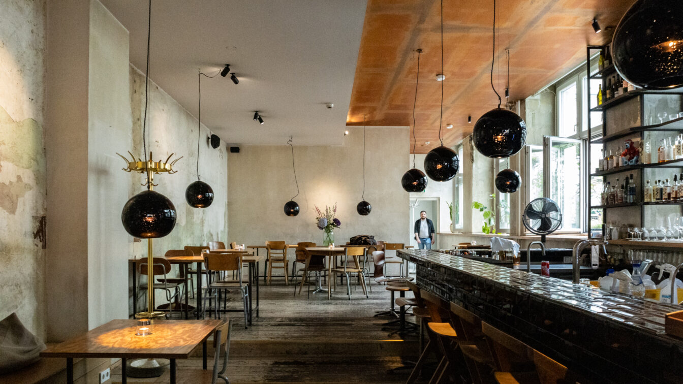 Innenansicht des stilvollen Michelberger Hotels in Berlin: Das Restaurant oder die Bar besticht durch modernes, urbanes Design mit rustikalem Holzboden, schwarzen Hängelampen, offener Bar und kreativem Mix aus Industrial- und Vintage-Elementen. Eine entspannte Atmosphäre für Gäste mit Sinn für Design und Genuss.