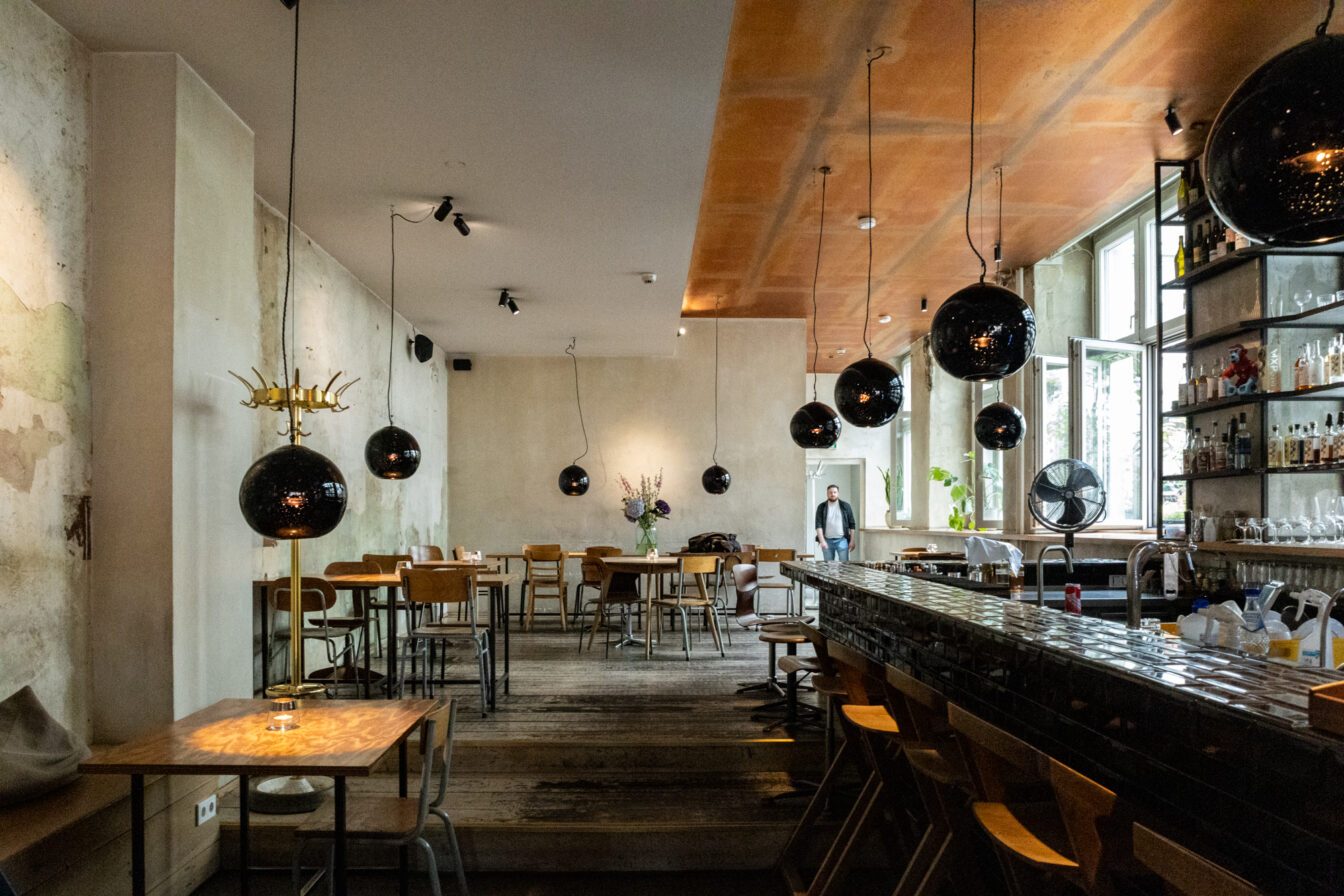 Innenansicht des stilvollen Michelberger Hotels in Berlin: Das Restaurant oder die Bar besticht durch modernes, urbanes Design mit rustikalem Holzboden, schwarzen Hängelampen, offener Bar und kreativem Mix aus Industrial- und Vintage-Elementen. Eine entspannte Atmosphäre für Gäste mit Sinn für Design und Genuss.