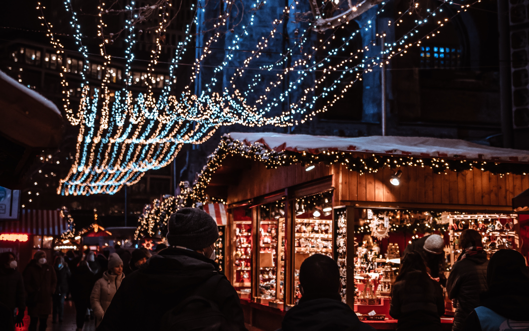 Menschen schlendern über einen festlich beleuchteten Weihnachtsmarkt in Berlin. Holzhütten mit Lichtern und Dekorationen bieten weihnachtliche Waren an, während über den Köpfen funkelnde Lichterketten den dunklen Abend erhellen.