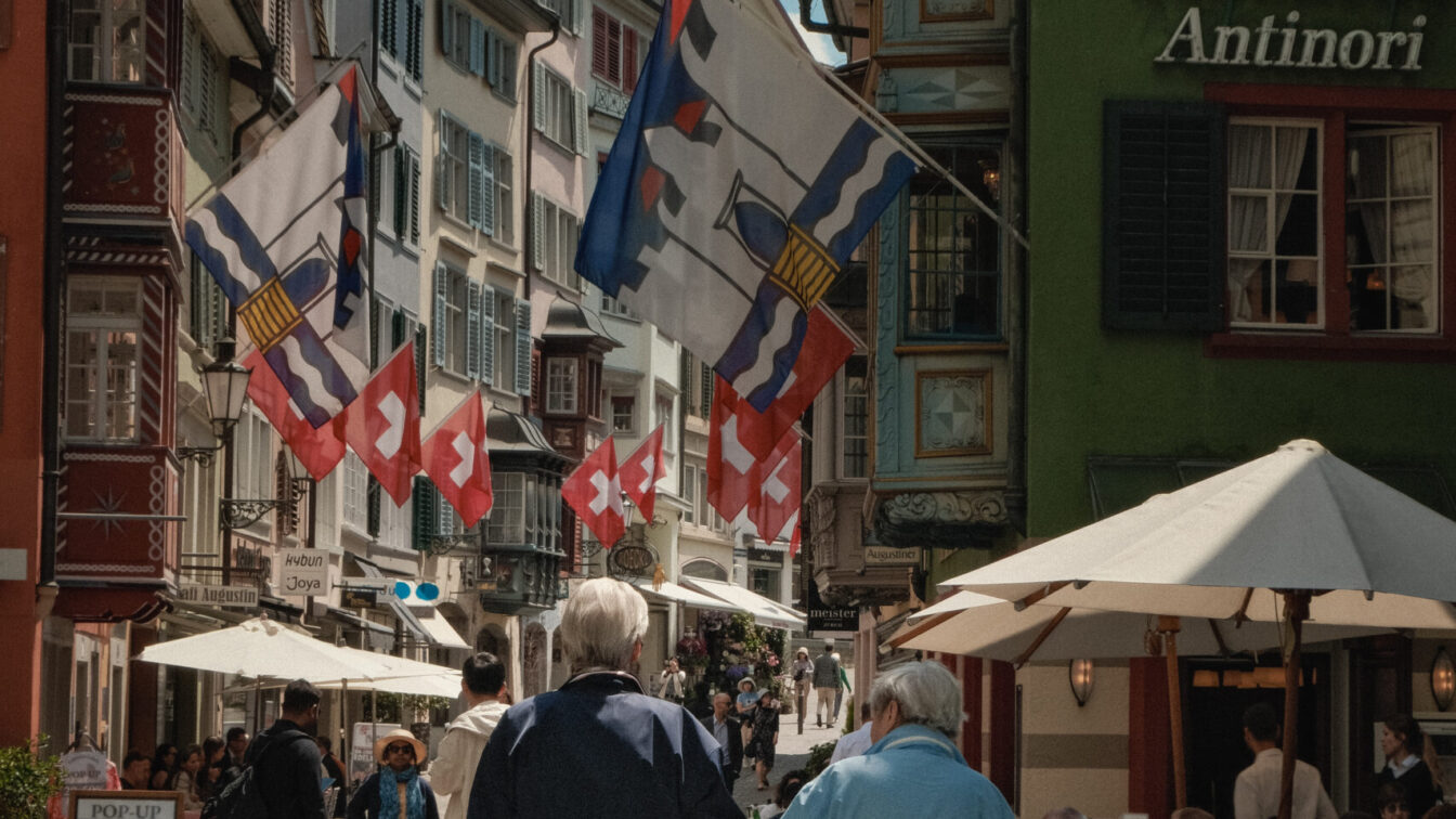 Zwei ältere Personen gehen Hand in Hand durch die Augustinergasse in Zürich, flankiert von historischen Häusern mit bunten Fassaden und Schweizer Fahnen. Vor den Restaurants sitzen Gäste an Tischen unter Sonnenschirmen, während die Straße lebendig wirkt